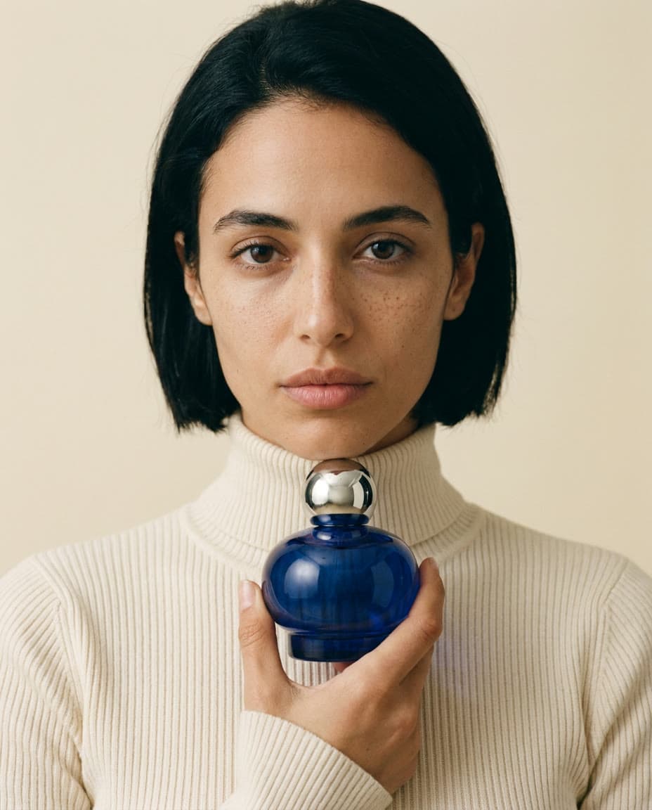 The same woman in a cream sweater, holding a cobalt perfume bottle to her collarbone