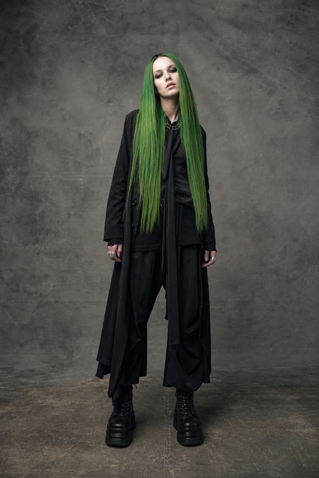 A woman with long green hair in a black coat against a grey backdrop